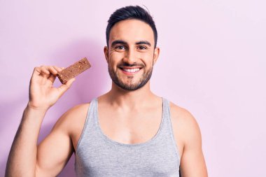 Sakallı, sakallı, enerji proteini yiyen, pembe arka planda mutlu ve pozitif görünen, kendine güvenen bir gülümsemeyle dişlerini gösteren genç bir adam.