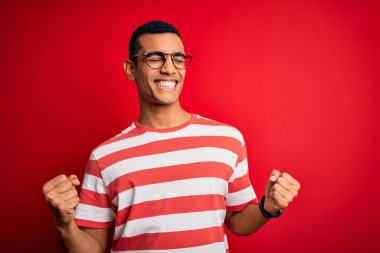 Genç ve yakışıklı Afro-Amerikan erkeği, sıradan çizgili tişört ve gözlük takıyor. Çok mutlu ve heyecanlı. Kazanan jestler yapıyor. Kutlama konsepti.