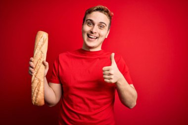 Genç ve yakışıklı kızıl saçlı adam izole edilmiş kırmızı arka planda taze ev yapımı ekmek tutuyor mutlu ve pozitif gülümsüyor, başparmağını kaldırıp mükemmel ve onay işareti yapıyor.