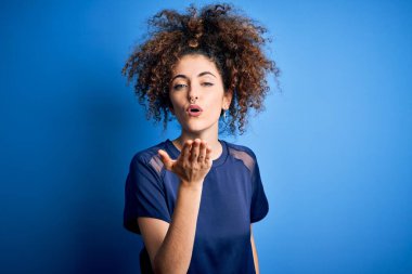 Kıvırcık saçlı, piercingli, sıradan mavi tişörtlü, kameraya bakıp havada öpücükler saçan, güzel ve seksi bir kadın. Aşk ifadesi.