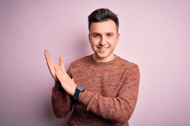 Pembe izole arka plan üzerine sıradan bir kazak giyen genç beyaz adam alkışlıyor ve mutlu ve neşeli, gururlu elleri alkışlıyor.