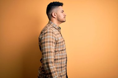 Genç, yakışıklı bir adam, günlük gömleğini giymiş, izole edilmiş sarı arka plan üzerinde duruyor, yan tarafa bakıyor, rahat bir görünüm takınıyor, doğal bir yüzü ve kendine güvenen bir gülümsemesi var..