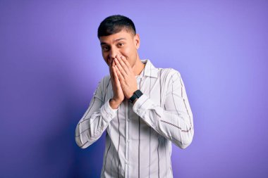 Zarif iş gömleği giyen genç yakışıklı İspanyol adam mor arka planda duruyor gülüyor ve utanıyor kıkırdıyor ağızları ellerle, dedikoduyla ve skandallarla kaplıyor.