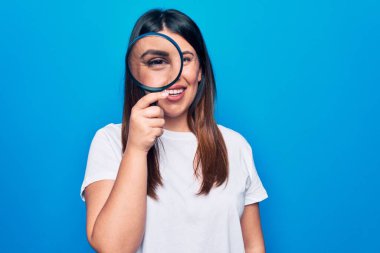 Genç, güzel esmer kadın mavi arka plan üzerinde büyüteç kullanıyor. Pozitif ve mutlu görünüyor. Kendine güvenen bir gülümsemeyle dişlerini gösteriyor.