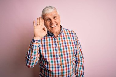 Kıdemli yakışıklı, beyaz gömlekli, soyutlanmış pembe arka planda. Mutlu ve güler yüzlü, dostça bir jest yapıyor.