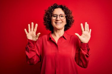Orta yaşlı güzel kıvırcık saçlı kadın kırmızı arka planda günlük gömlek ve gözlük takıyor ve parmaklarıyla on numarayı gösterirken kendinden emin ve mutlu gülümsüyor..