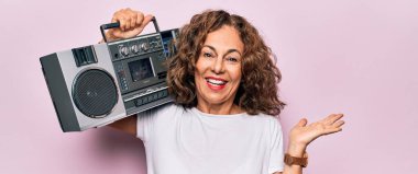 Orta yaşlı esmer hippi kadın pembe izole edilmiş arka planda retro müzik teybi tutuyor. Mutlu bir gülümsemeyle başarıyı kutluyor ve kazanan yüz ifadesini kaldırarak elini kaldırıyor.