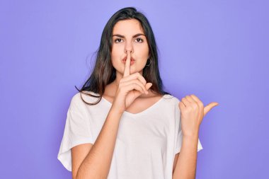 Genç, güzel, esmer bir kadın, mor arka plan üzerine gündelik beyaz tişört giyip sessiz olmak istiyor. Parmakları dudaklarında, elleri yandan göstererek. Sessizlik ve gizli konsept.