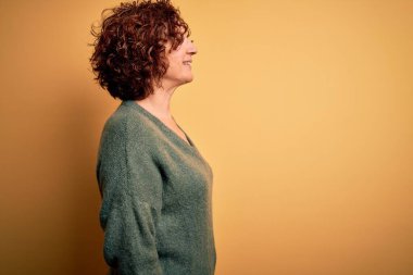 Orta yaşlı, kıvırcık saçlı, sıradan bir süveter giyen, yalıtılmış sarı arka plan giyen, rahat profil pozu veren, kendine güvenen bir gülümsemesi olan güzel bir kadın..