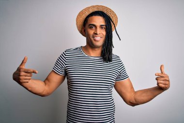Afrika kökenli Amerikalı genç bir adam tatilde. Çizgili tişört ve şapka giyiyor. Kendine güvenen, gülümseyen, gururlu ve mutlu birini işaret ediyor..