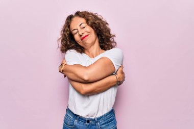Middle age beautiful woman wearing casual t-shirt standing over isolated pink background hugging oneself happy and positive, smiling confident. Self love and self care clipart