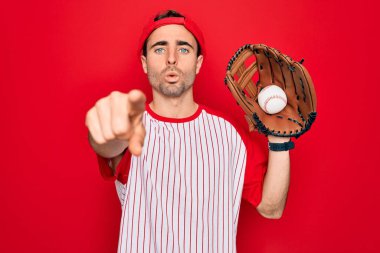 Mavi gözlü genç, yakışıklı, beysbol oynayan, eldiven ve topla kamerayı işaret eden, el işareti, ön taraftan pozitif ve kendinden emin bir jest yapan.