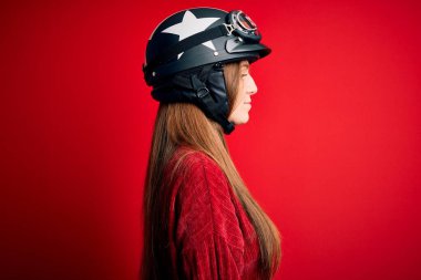Kırmızı arka planda moto miğfer takan güzel, kızıl saçlı, güzel bir motosikletçi kadın. Yan tarafa bakıyor, rahat poz veriyor, kendine güvenen bir gülümsemeyle doğal bir yüz ifadesi sergiliyor..