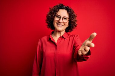 Orta yaşlı, kıvırcık saçlı, rahat gömlekli, kırmızı arka planda gözlüklü neşeli gülümsemeli, yardım eli uzatan ve kabul gören bir kadın..