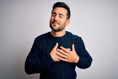 Sakallı, sıradan bir süveter giyen, beyaz arka plan üzerinde duran, elleri göğsünde, gözleri kapalı ve yüzünde minnet ifadesi olan genç yakışıklı bir adam. Sağlık kavramı.