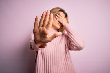 Genç, güzel, sarışın bir kadın, izole edilmiş arka planda pembe süveter giyiyor, gözleri ellerle örtüyor ve üzgün ve korku ifadeleriyle jest yapıyor. Utanç verici ve olumsuz bir kavram..
