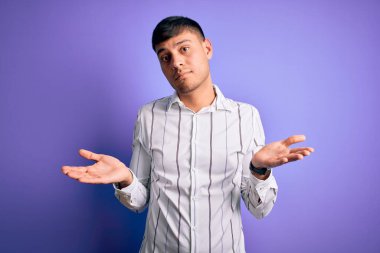 Zarif bir iş gömleği giyen yakışıklı genç adam mor arka planda duruyor. Kolları açık ve kafası karışık, hiçbir fikri yok..