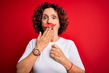 Orta yaşlı kıvırcık saçlı kadının kırmızı arka planda komik bıyıkları var. Ağzı kapalı. Utanç verici bir şekilde ağzı kapalı.