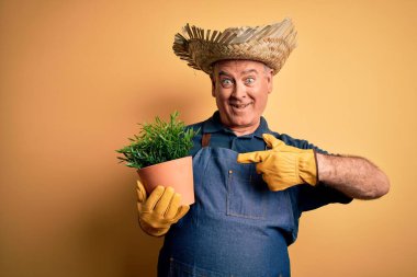 Sarı arka planda önlük ve şapka takan orta yaşlı çiftçi elini ve parmağını göstererek mutlu bir şekilde bitki saksısı tutuyor.