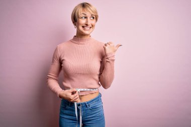 Kısa saçlı, zayıf, genç bir kadın. Ölçüm bandı kullanarak, gülümseyen yüzüyle başparmağıyla işaret ediyor.