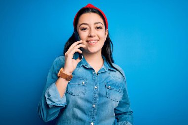 Genç esmer kadın mavi izole edilmiş arka planda akıllı telefondan konuşuyor. Mutlu bir yüzü var. Kendine güvenen bir gülümsemeyle dişlerini gösteriyor.