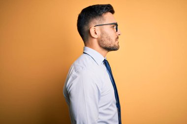 Sarı arka plan üzerinde duran kravat ve gözlük takan genç yakışıklı işadamı yan yana bakıyor, rahat poz veriyor ve kendine güvenen bir gülümseme takınıyor..