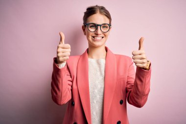 Ceket ve gözlük takan genç, güzel bir kızıl kadın pembe arka planda olumlu bir jest yaptığını onaylıyor. Elleriyle gülümsüyor ve başarı için seviniyor. Kazanan jest.