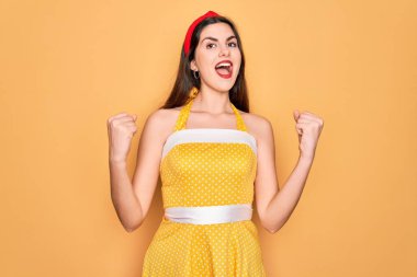 Sarı arka plan üzerine 50 'li yılların moda elbisesi giymiş genç ve güzel bir kadın kollarını kaldırıp, gözleri açık bir şekilde başarıyı kutluyor ve şaşırıyor. Kazanan konsept.