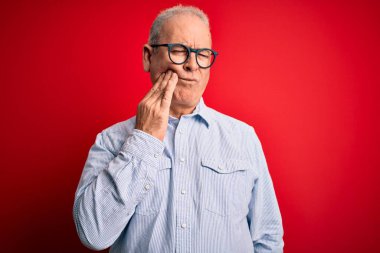 Orta yaşlı, beyaz, sıradan bir gömlek ve kırmızı arka planda gözlük takan yakışıklı bir adam diş ağrısı ya da diş ağrısı yüzünden ağız ve acı dolu bir ifadeyle birbirine dokunuyor. Dişçi