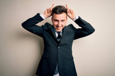 Zarif takım elbiseli ve kravatlı genç yakışıklı iş adamı izole arka planda komik ve çılgın pozlar veriyor parmakları kafasında tavşan kulağı, neşeli bir gülümseme