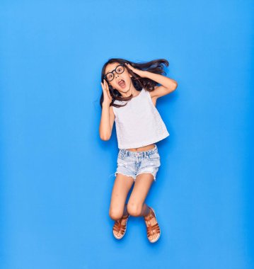 Günlük kıyafetler ve gözlük takan sevimli İspanyol kız ağzı açık kalınca şaşırdı. İzole edilmiş mavi arka planda eller havada zıplıyor.