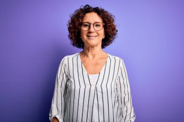 Orta yaşlı güzel kıvırcık saçlı kadın izole edilmiş arka planda sıradan çizgili gömlek giyiyor ve yüzünde mutlu ve havalı bir gülümseme var. Şanslı kişi..