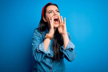 Genç esmer kadın, mavi izole edilmiş arka plan üzerine sıradan bir kot gömlek giyip elleri ağzında kızgın bir şekilde bağırıyor.