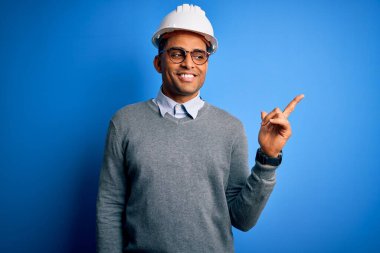 Eli ve parmağı kameraya bakan, yüzünde kocaman bir gülümsemeyle, güvenlik kaskı takan, Afrika asıllı, genç, Amerikalı mühendis..