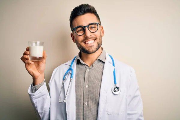幸せな顔をして立っていると歯を示す自信を持って笑顔で笑みを浮かべて孤立した背景にミルクのガラスを保持聴診器を身に着けている若い医師 — ストック写真