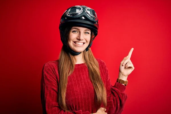 カメラを見ている側に手と指で指して 顔に大きな笑顔で赤い背景に元ヘルメットを身に着けている若い美しい赤毛の元サイクリストの女性 — ストック写真