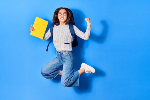 Young beautiful chinese student girl wearing glasses and backpack smiling happy. Jumping with smile on face holding book over isolated blue background