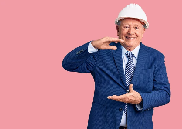 Senior handsome grey-haired man wearing suit and architect hardhat gesturing with hands showing big and large size sign, measure symbol. smiling looking at the camera. measuring concept.