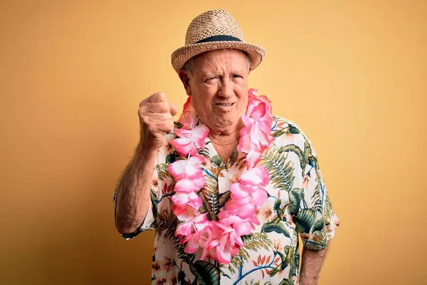 Grey haired senior man wearing summer hat and hawaiian lei over yellow background angry and mad raising fist frustrated and furious while shouting with anger. Rage and aggressive concept.