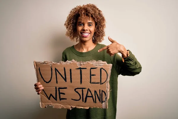 African american activist woman asking for unity holding banner with united stand message with surprise face pointing finger to himself