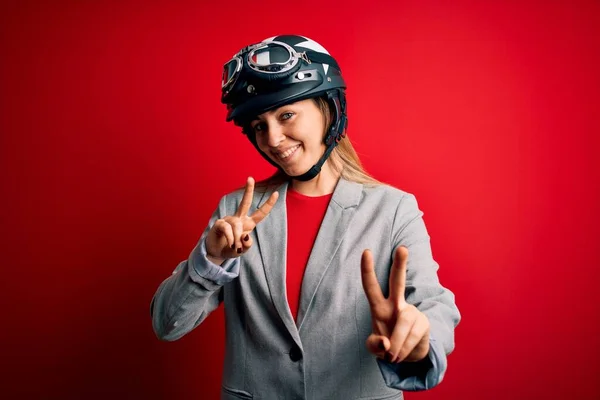 年轻美丽的金发女摩托车手 戴着摩托车头盔 头戴红色背景 微笑着看着摄像机 手指头在做胜利的标志 第二点 — 图库照片