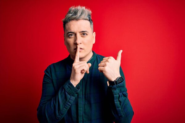 Young handsome modern man wearing elegant green shirt over red isolated background asking to be quiet with finger on lips pointing with hand to the side. Silence and secret concept.