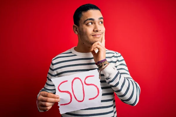 Young brazilian man with problem holding banner with sos message over red background serious face thinking about question, very confused idea