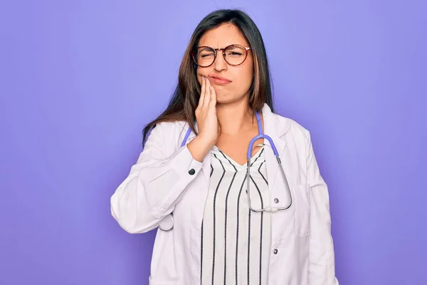 歯の痛みや歯の病気のために痛みを伴う式で手で口に触れる紫色の背景に聴診器や医療用コートを身に着けている専門医の女性 歯医者 — ストック写真