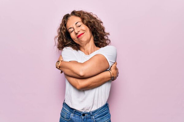 Middle age beautiful woman wearing casual t-shirt standing over isolated pink background hugging oneself happy and positive, smiling confident. Self love and self care
