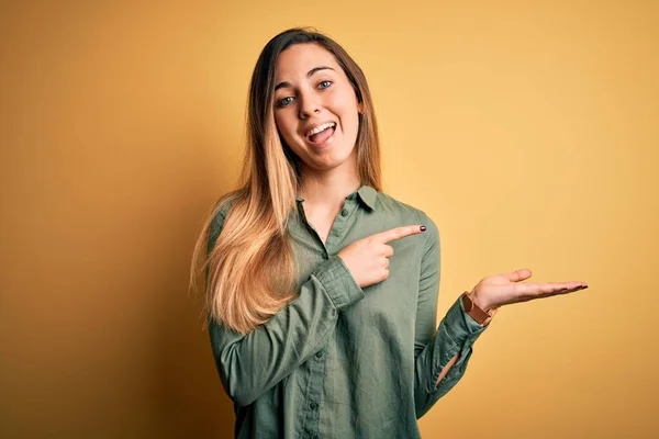 年轻美丽的金发女子 蓝眼睛 身穿绿色衬衫 背景黄色 手牵着手 指指点点着摄像机 惊讶地微笑着 — 图库照片