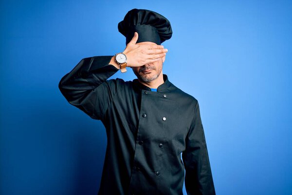 Young handsome chef man wearing cooker uniform and hat over isolated blue background covering eyes with hand, looking serious and sad. Sightless, hiding and rejection concept