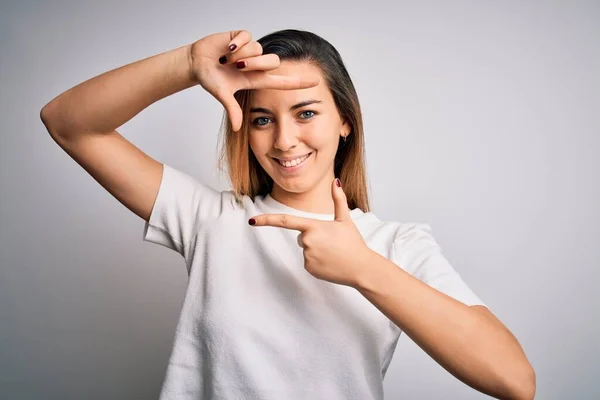 白い背景にカジュアルなTシャツを着た青い目の若い美しいブロンドの女性は 幸せな顔をした手と指でフレームを作り笑顔 創造性と写真の概念 — ストック写真