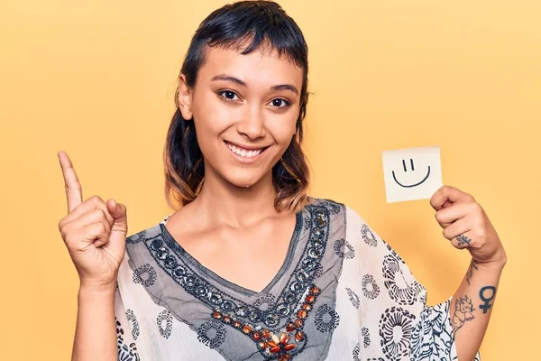 Young woman holding smile emoji reminder smiling happy pointing with hand and finger to the side
