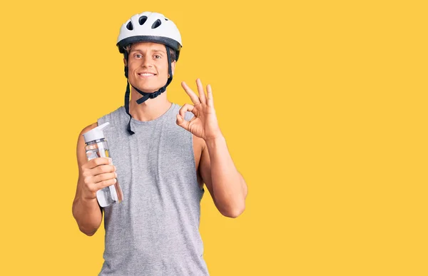 Young handsome man wearing bike helmet holding bottle of water doing ok sign with fingers, smiling friendly gesturing excellent symbol